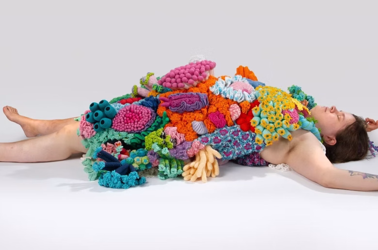 A person laying on the ground is covered in sculptures that appear like colorful coral.